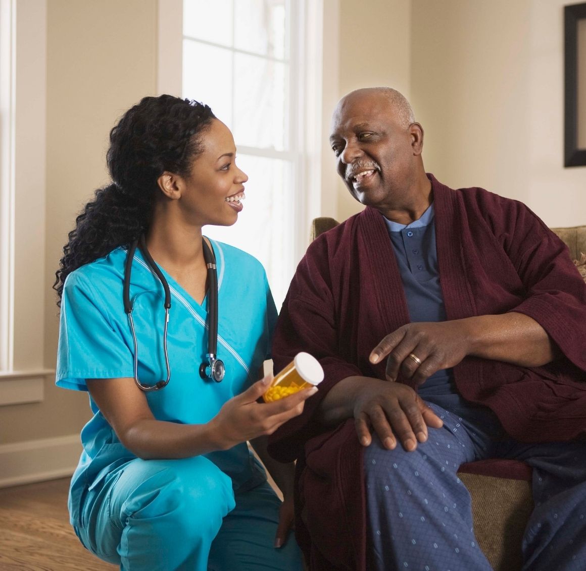 Medication Management Assistance Caregiver in scrubs helping an elderly man review medication bottles while sitting together in a well lit living room.