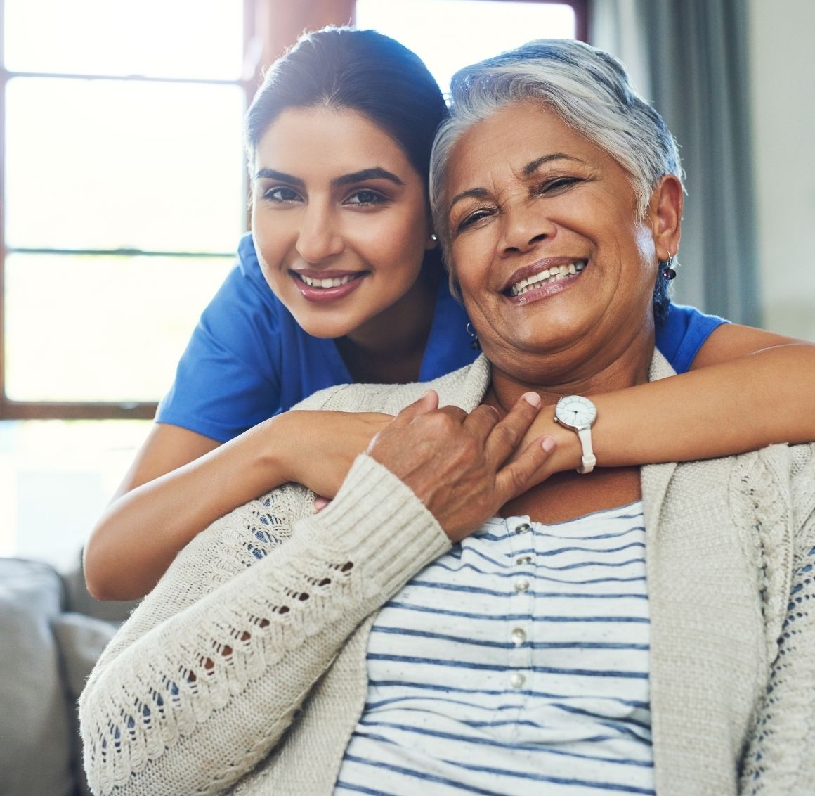 Happy caregiver wrapping her arms around an elderly woman, both smiling warmly in a home environment.