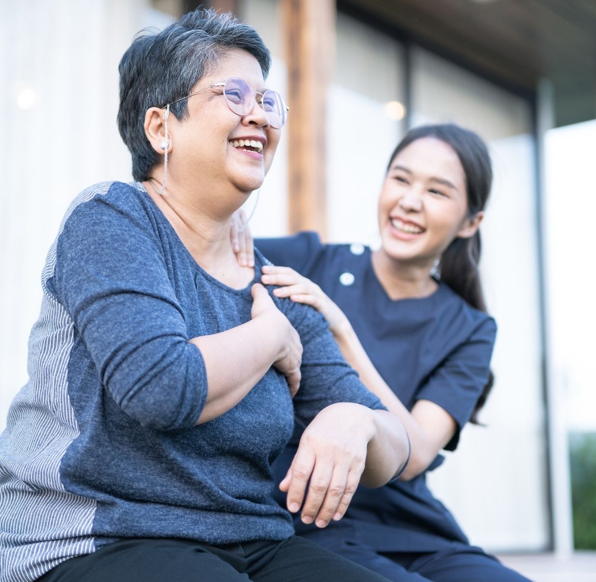 Caregiver providing companionship to elderly woman outdoors Caregiver smiling and touching the shoulder of an elderly woman as they sit outside and share a joyful moment.