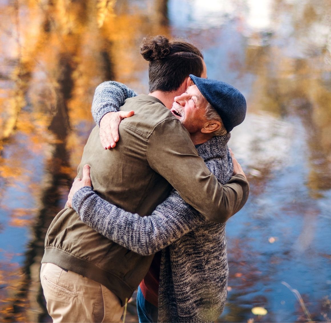 Elderly man hugging caregiver outdoors in park Elderly man warmly hugging his caregiver in an outdoor park setting with autumn trees and water in the background.
