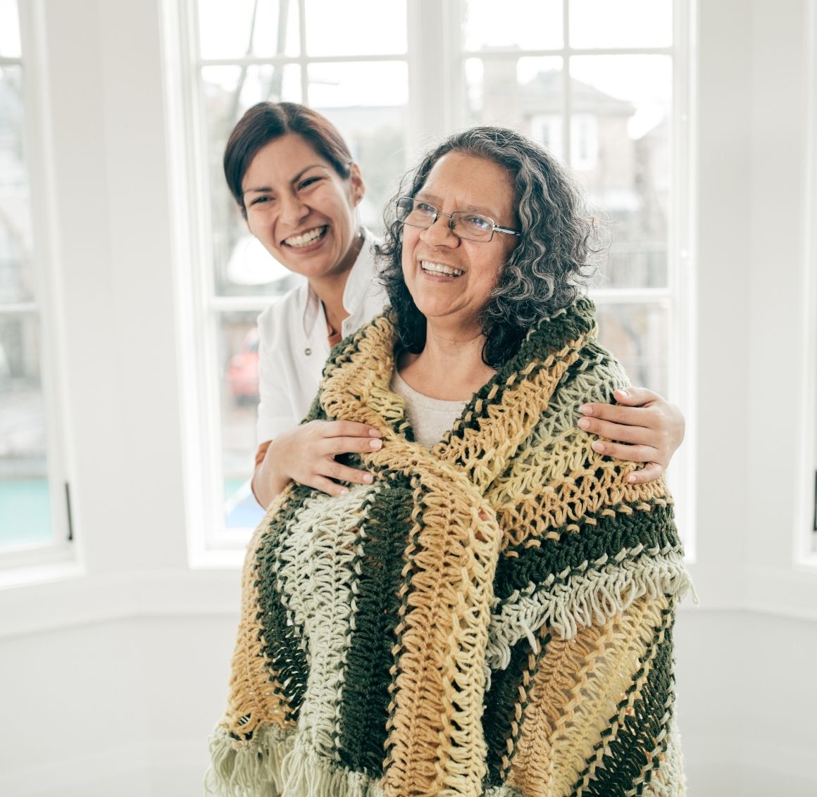 Respite Care Assistance with Caregiver Providing Comfort Caregiver wrapping a warm knitted blanket around a smiling elderly woman near bright windows symbolizing comfort and emotional support.