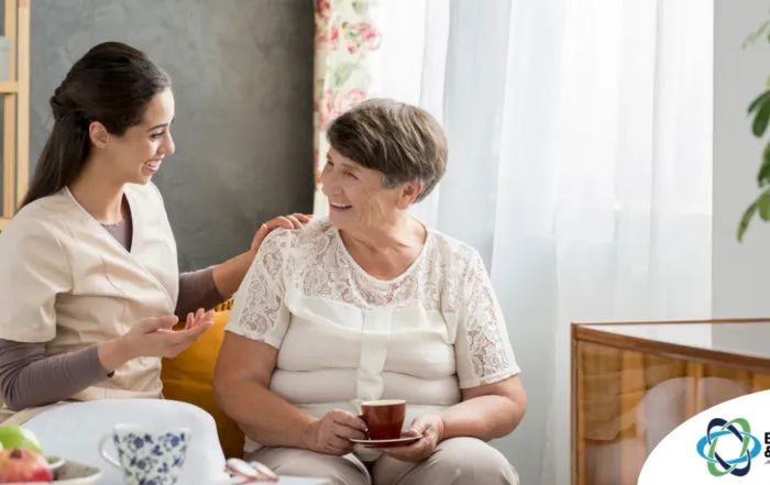 Smiling caregiver with her hand on an older woman’s shoulder as they share tea on the couch, showing home care vs assisted living