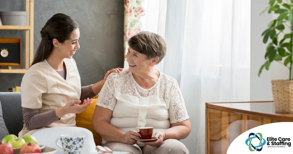 Smiling caregiver with her hand on an older woman’s shoulder as they share tea on the couch, showing home care vs assisted living